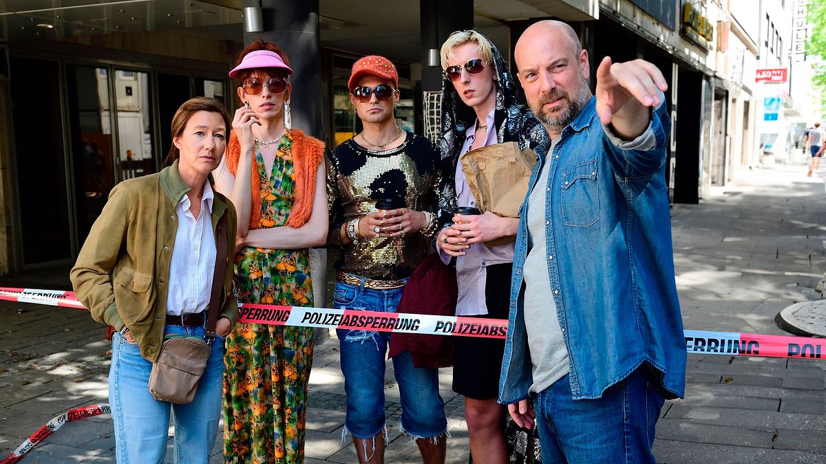 „Polizeiruf 110: Ein feiner Tag für den Bananenfisch“ läuft am 18. Mai. Schaulustige haben sich am Tatort versammelt. Kommissar Dennis Eden zeigt vor sich, die Schaulustigen und Kommissarin Cris Blohm blicken in die Ferne.