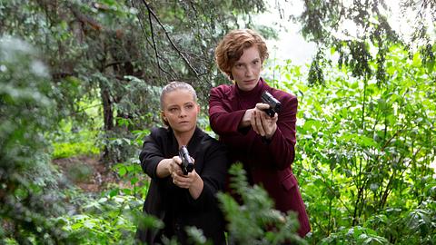 Jasna Fritzi Bauer und Luise Wolfram stehen in einem Wald und zielen mit ihren Schusswaffen. Bauer trägt schwarz und Wolfram rot.  - Foto:  Radio Bremen/Claudia Konerding
