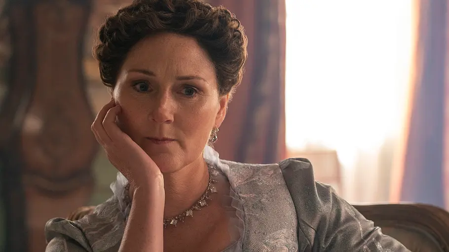 Violet Bridgerton Spin off Violet Bridgerton mit hochgestecktem Haar und silbergrauem Kleid sitzt auf einem Stuhl und stützt ihre Wange auf die Hand.