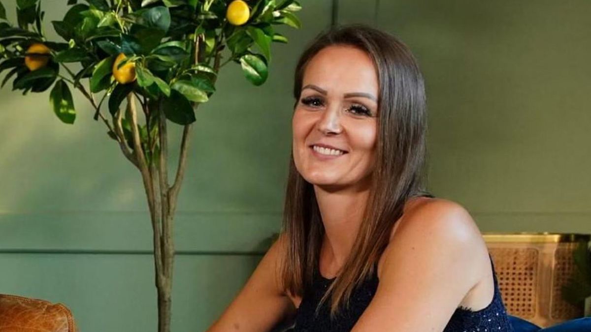 Stefanie Hausen Stefanie lächelt in die Kamera. Sie sitzt vor einen grünen Wand und neben einem kleinen Zitronenbaum