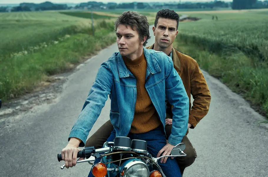 Marc De Vuyst (Tijmen Govaerts, l.) und Franky Verhellen (Aimé Claeys, r.) sitzen hintereinander auf einem Motorrad auf einer schmalen Straße zwischen grünen Feldern.
