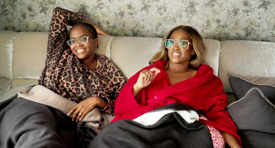 Motsi Oti Mabuse Motsi und Oti Mabuse sitzen mit Brillen und Decken auf einem Sofa, eine im Leopardenoberteil, die andere im roten Bademantel.