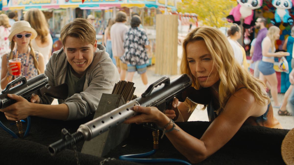 Connie Nielsen hält auf einem Jahrmarkt ein Gewehr in der Hand und spielt die Rolle der Becca in ‚Nobody 2‘. Neben ihr steht ihr Sohn, gespielt von Gage Munroe, ebenfalls mit einem Spielgewehr in der Hand.
