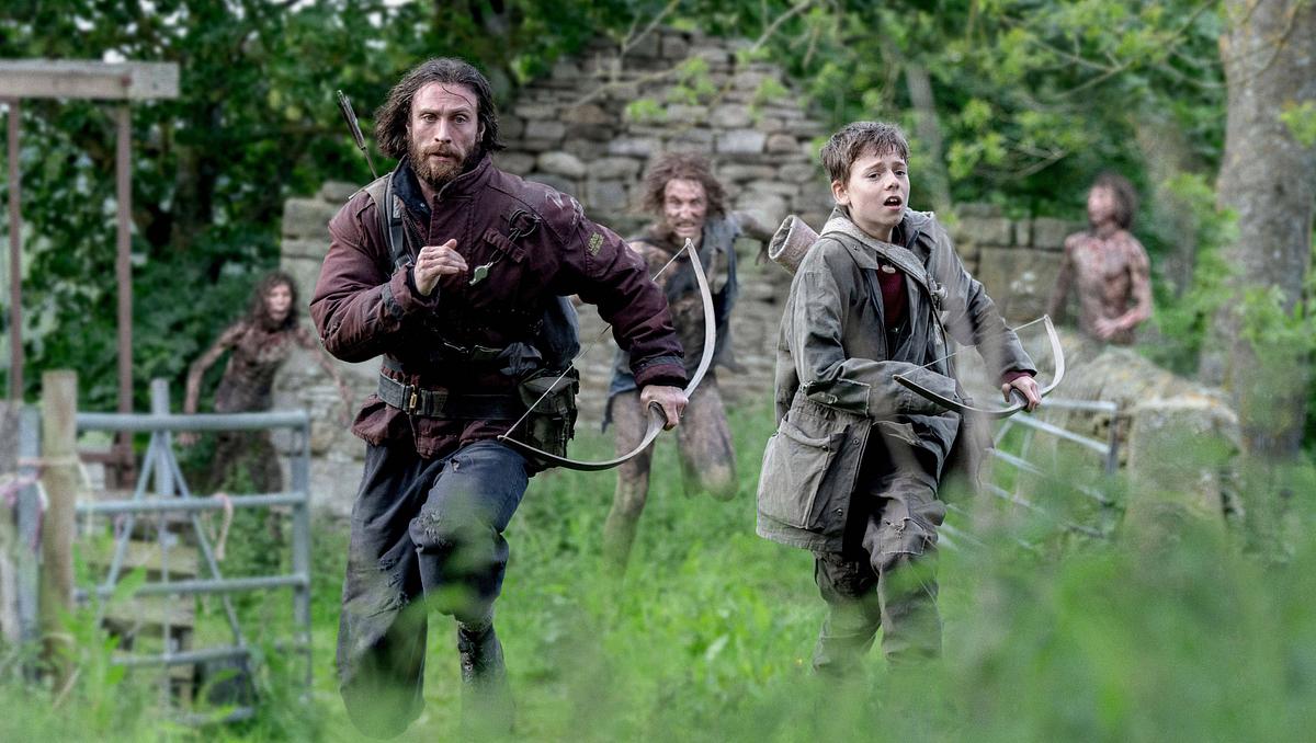 28 Years Later mit Cillian Murphy Man sieht Aaron Taylor-Johnson und Alife WIlliams, wie sie mit Pfeil und Bogen vor drei Untoten in einer Graslandschaft mit den Überresten eines Gebäudes, sowie einem Zaun weglaufen.