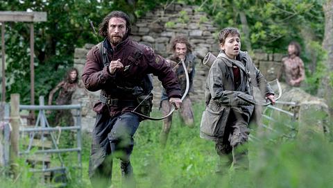 Man sieht Aaron Taylor-Johnson und Alife WIlliams, wie sie mit Pfeil und Bogen vor drei Untoten in einer Graslandschaft mit den Überresten eines Gebäudes, sowie einem Zaun weglaufen. - Foto: IMAGO / Landmark Media