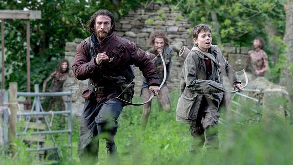 Man sieht Aaron Taylor-Johnson und Alife WIlliams, wie sie mit Pfeil und Bogen vor drei Untoten in einer Graslandschaft mit den Überresten eines Gebäudes, sowie einem Zaun weglaufen. - Foto: IMAGO / Landmark Media