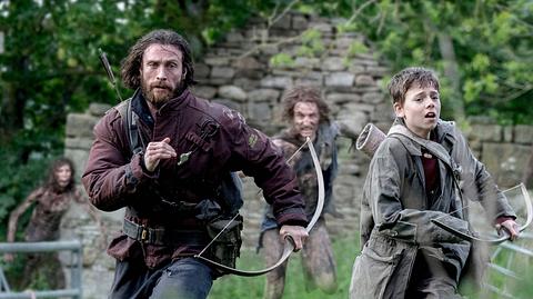 Jamie (Aaron Taylor-Johnson, links) und Spike (Alfie Williams) laufen auf einer grünen Wiese vor einer Ruine vor Infizierten weg. Sie tragen Funktionskleidung und sind mit Pfeil und Bogen bewaffnet. Die Infizierten tragen keine sonderlich auffällige Kleidung, aber schauen finster drein, während Spike und Jamie eher ernst davonlaufen. - Foto:  IMAGO / Landmark Media
