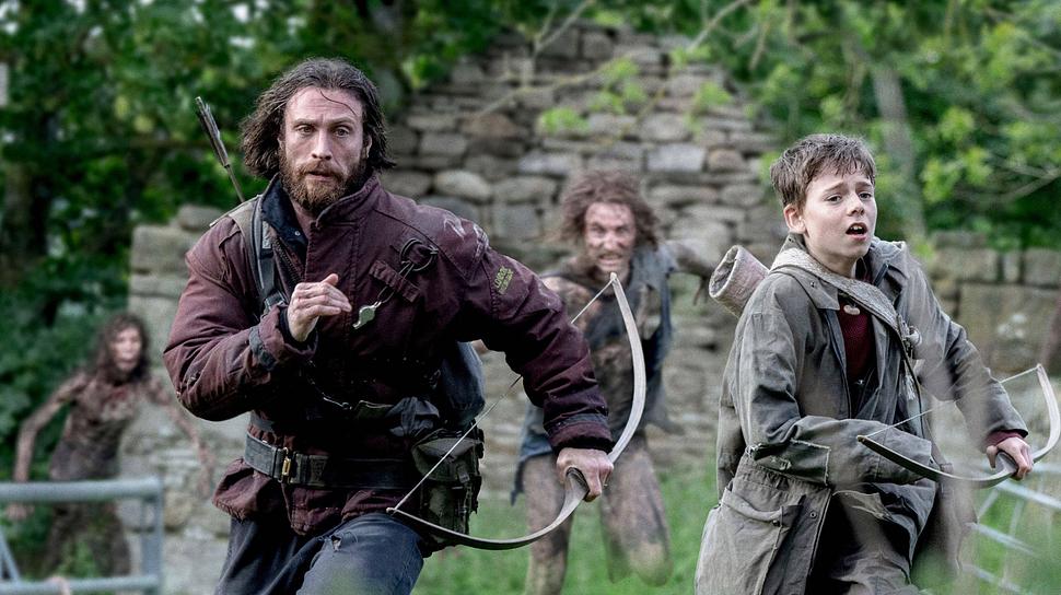 Jamie (Aaron Taylor-Johnson, links) und Spike (Alfie Williams) laufen auf einer grünen Wiese vor einer Ruine vor Infizierten weg. Sie tragen Funktionskleidung und sind mit Pfeil und Bogen bewaffnet. Die Infizierten tragen keine sonderlich auffällige Kleidung, aber schauen finster drein, während Spike und Jamie eher ernst davonlaufen. - Foto:  IMAGO / Landmark Media