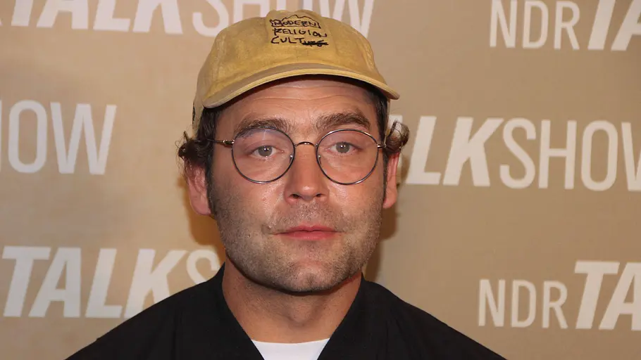 Bosse Aki Bosse bei einem NDR-Event: Der Musiker trägt Brille und Kappe sowie eine dunkle Jacke und blickt neutral in die Kamera vor einer „NDR Talk Show“-Wand