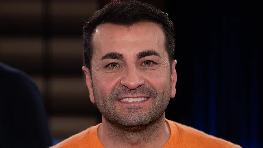 Porträt eines lächelnden Mannes mit dunklen Haaren und orangefarbenem Pullover vor dunklem Hintergrund