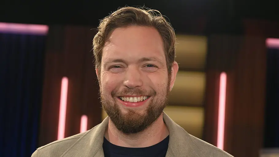 Bastian Bielendorfer Bastian Bielendorfer im TV-Studio: Der Comedian lächelt in die Kamera und trägt ein Sakko über einem dunklen Shirt, im Hintergrund sind Studiolichter zu sehen