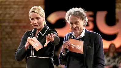 Judith Rakers und Giovanni di Lorenzo moderieren eine Talkrunde, beide halten Mikrofone und Moderationskarten in der Hand und sprechen auf einer Bühne vor Publikum - Foto: IMAGO / Future Image