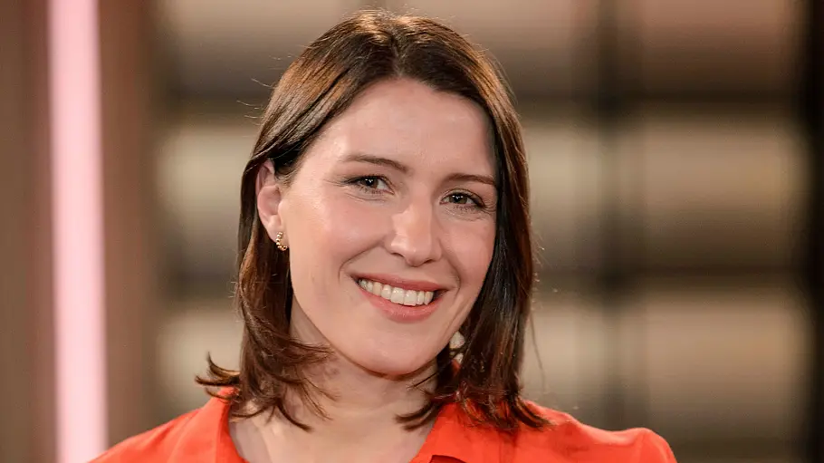 Porträt einer lächelnden Frau mit schulterlangem braunem Haar und roter Bluse in einem Studio mit unscharfem Hintergrund.