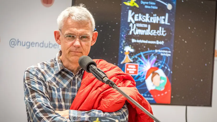Älterer Mann mit Brille und kariertem Hemd steht mit verschränkten Armen an einem Mikrofon, hinter ihm ein Bildschirm mit Buchcover und Veranstaltungslogo.
