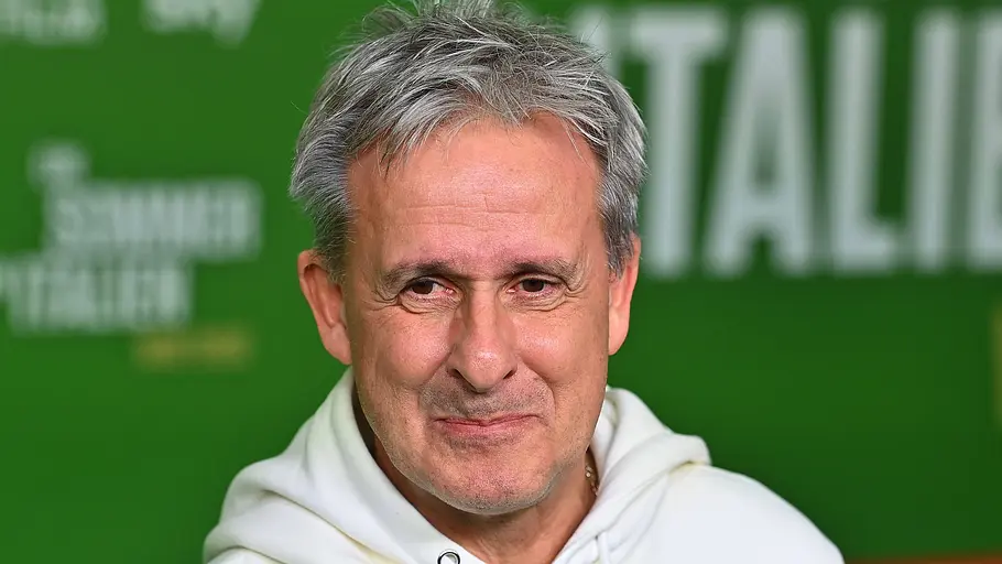 Porträt eines grauhaarigen Mannes in weißem Hoodie vor einer grünen Veranstaltungswand mit Schriftzug im Hintergrund.