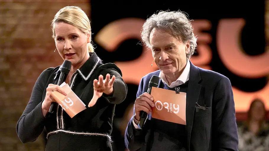 Judith Rakers und Giovanni di Lorenzo Judith Rakers und Giovanni di Lorenzo stehen mit Mikrofonen und orangefarbenen Karten vor Publikum. Judith streckt eine Hand nach vorn.