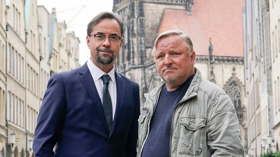 Axel Prahl und Jan Josef Liefers Jan Josef Liefers im blauen Anzug und Axel Prahl in grauer Jacke stehen auf einer Straße vor einer Kirche.