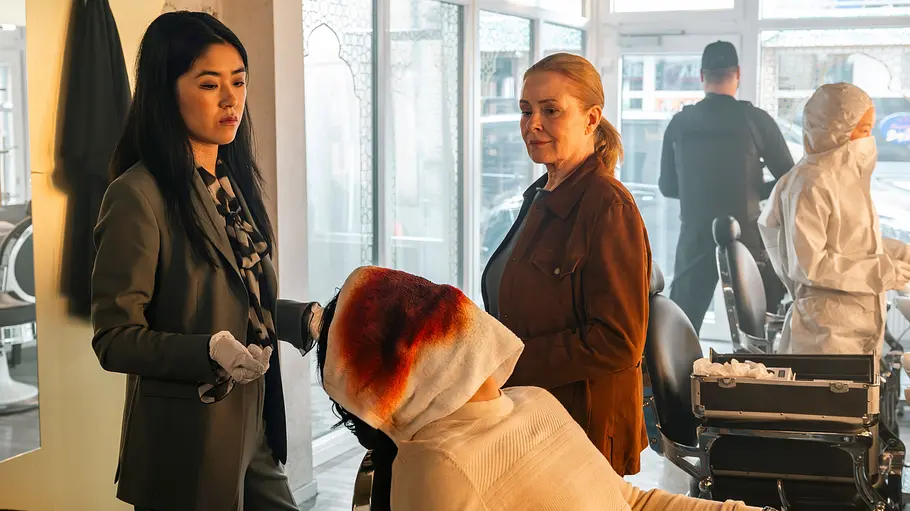 Dr. Clara Chu und Anne Fuchs stehen in einem Barbershop vor einer ermordeten Person mit blutigem Handtuch über dem Kopf, während die Spurensicherung im Hintergrund arbeitet.