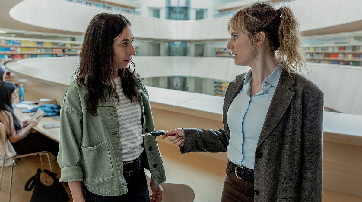 Tatort Rapunzel Zürich Man sieht die Züricher Ermittlerinnen Ott (links) und Grandjean (rechts). Sie stehen in einer modernen Bibliothek, die wie mehrere Kreise übereinander geschichtet ist. Ott trägt eine grüne Jacke und ein weiß-schwarz gestreiftes Oberteil, sowie eine schwarze Hose. Eine schwarze Hose trägt auch Grandjeans plus eine hellblaue Bluse und ein graues Jacket.