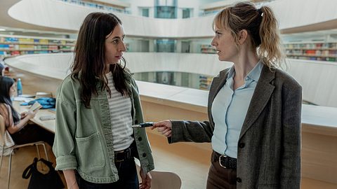 Man sieht die Züricher Ermittlerinnen Ott (links) und Grandjean (rechts). Sie stehen in einer modernen Bibliothek, die wie mehrere Kreise übereinander geschichtet ist. Ott trägt eine grüne Jacke und ein weiß-schwarz gestreiftes Oberteil, sowie eine schwarze Hose. Eine schwarze Hose trägt auch Grandjeans plus eine hellblaue Bluse und ein graues Jacket. - Foto: SRF/Sava Hlavacek