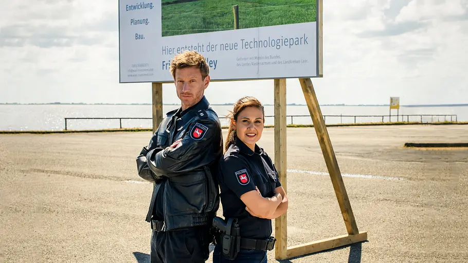 Die digitale Zukunft beginnt in Leer mit dem Friesland-Valley-Technologiepark. Süher Özlügül (Sophie Dal) und Henk Cassens (Maxim Mehmet) fühlen sich mit dem Auftrag der Begleiteskorte für den Investmentmanager nicht wertgeschätzt als Polizisten. Friesland - Haifischbecken: Henk Cassens (Maxim Mehmet) und Süher Özlügül (Sophie Dal) stehen Rücken an Rücken vor einer Plakatwand die einen Technologiepark ankündigt.