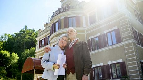Hildegard und Alfons stehen vor dem Fürstenhof. Er hält sie im Arm. Sie hat einen Großbrief in der Hand - Foto: ARD/Christof Arnold