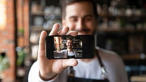 Mann hält kleines Smartphone für Selfie lächelnd vor sich - Foto: iStock/fizkes