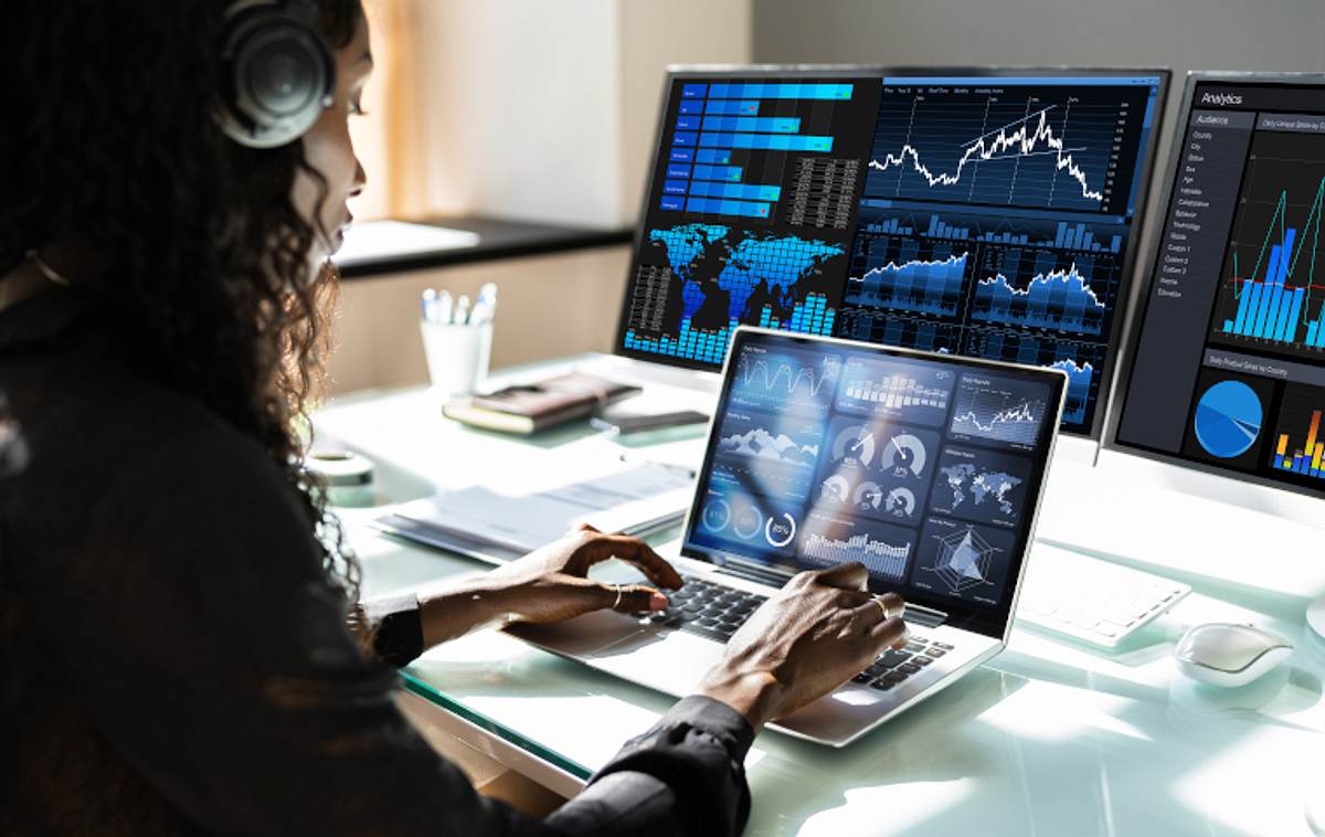 Frau arbeitet an Laptop und hat zwei externe Bildschirme angeschlossen. Alle Displays zeigen Statistiken an.