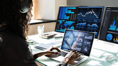 Frau arbeitet an Laptop und hat zwei externe Bildschirme angeschlossen. Alle Displays zeigen Statistiken an. - Foto: iStock/AndreyPopov