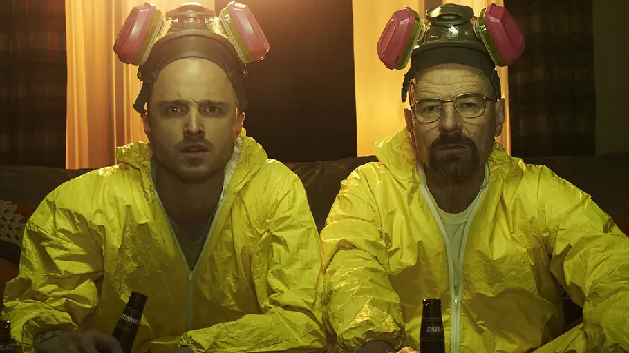Walter White und Jesse Pinkman, die beiden Hauptdarsteller der Serie „Breaking Bad“ von Vince Gilligan, sitzen mit Bierflaschen auf dem Sofa und tragen Schutzanzüge.