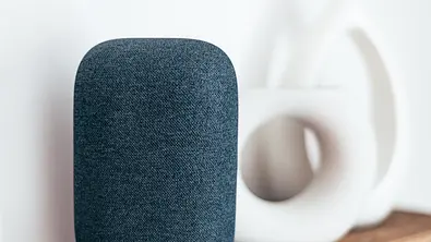 Ein intelligenter Lautsprecher in Grau auf einem Holzregal, daneben dekorative Vasen - Foto: iStock/Manuta