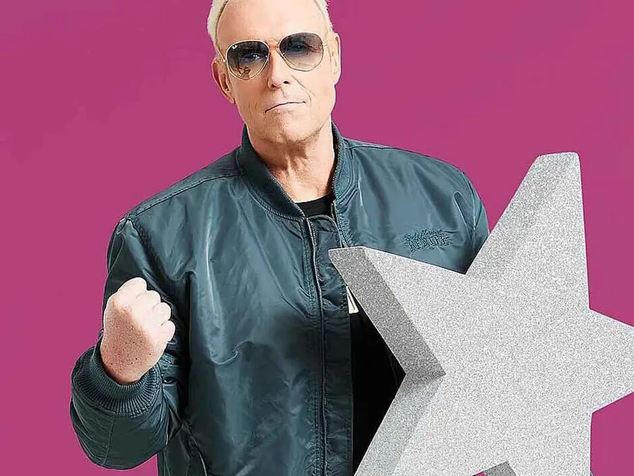 Promi Big Brother-Gewinner 2022: Rainer Gottwald Rainer Gottwald trägt Sonnenbrille und dunkle Jacke, hebt eine Faust und hält einen großen silbernen Stern vor pinkem Hintergrund.