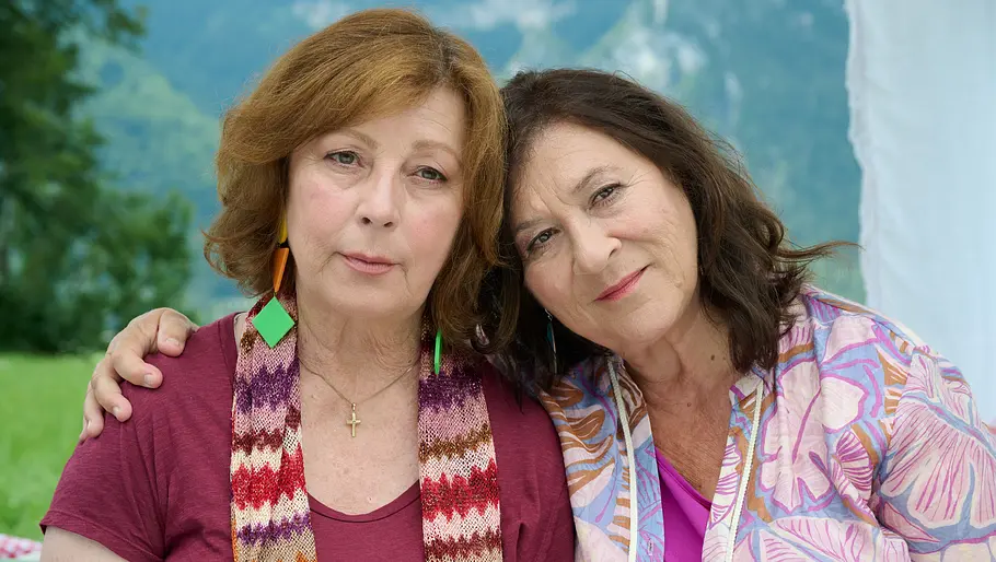 Eva Lorenz (Eva Mattes, r.) und ihre Schwester Liz White (Bettina Redlich, l.) Lena Lorenz - Schutzlos: Eva Lorenz (Eva Mattes) und Liz White (Bettina Redlich) sitzen mit melancholischem Blick nebeneinander. Eva hat den Arm um Liz gelegt und schmiegt ihren Kopf an den ihrer Schwester. Im Hintergrund liegt ein Bergpanorama.