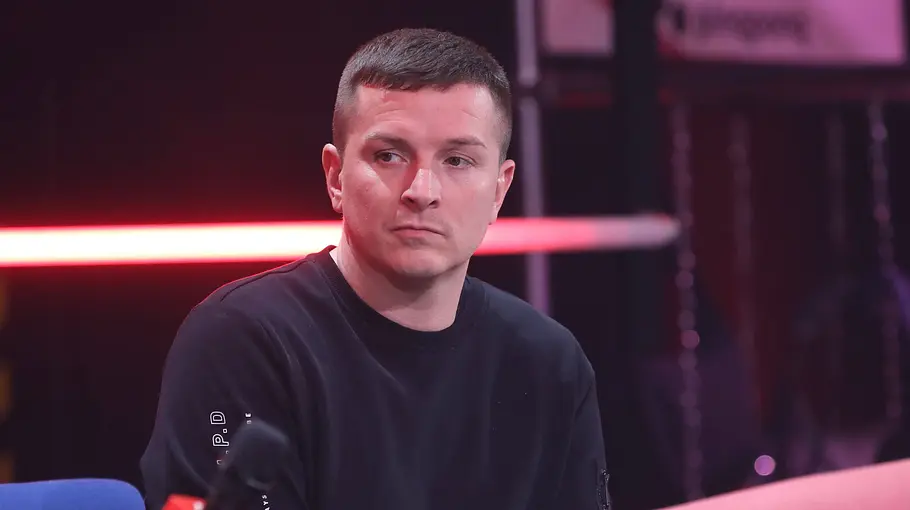 Aaron Troschke Promi BB Aus Aaron Troschke sitzt mit Mikrofon vor einer roten Lichtleiste. Auf niedrigen Tischen stehen Wassergläser.