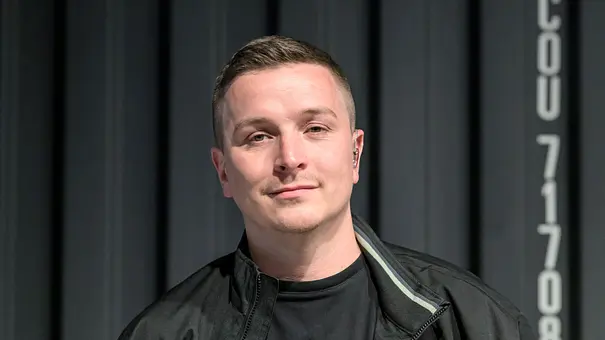 Aaron Troschke mit kurzen Haaren, schwarzer Jacke und schwarzem Shirt steht vor einer dunklen Metallwand mit weißer Beschriftung. - Foto: Imago/HMB-Media Aaron Troschke mit kurzen Haaren, schwarzer Jacke und schwarzem Shirt steht vor einer dunklen Metallwand mit weißer Beschriftung. - Foto: Imago/HMB-Media
