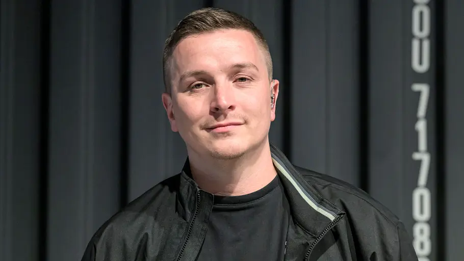 Aaron Troschke „Schlag den Aaron“. Aaron Troschke mit kurzen Haaren, schwarzer Jacke und schwarzem Shirt steht vor einer dunklen Metallwand mit weißer Beschriftung.