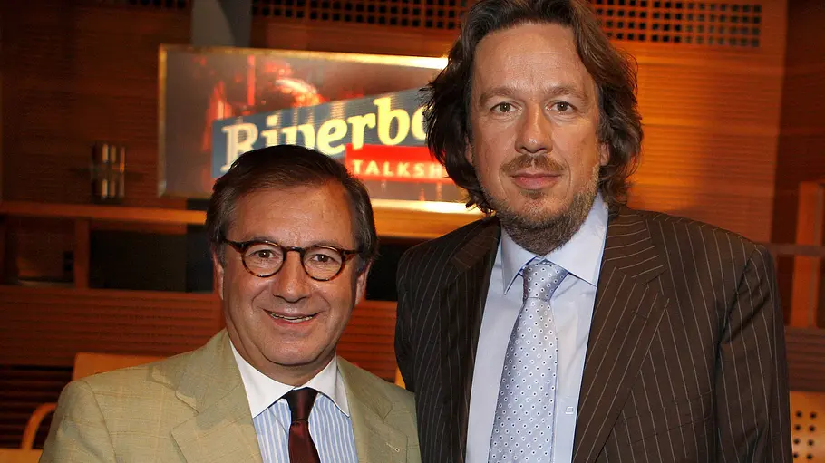 Jan Hofer und Jörg Kachelmann stehen in Anzügen und Krawatten nebeneinander vor einem „Riverboat“-Schild.
