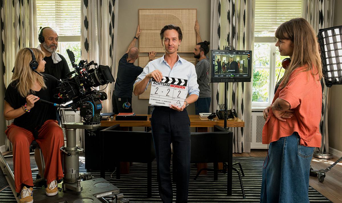 Man sieht Tom Schilling in einer schwarzen Hose und einem weißen Hemd. Er hält eine Filmklappe in der Hand und wird von einer Filmcrew umringt. 