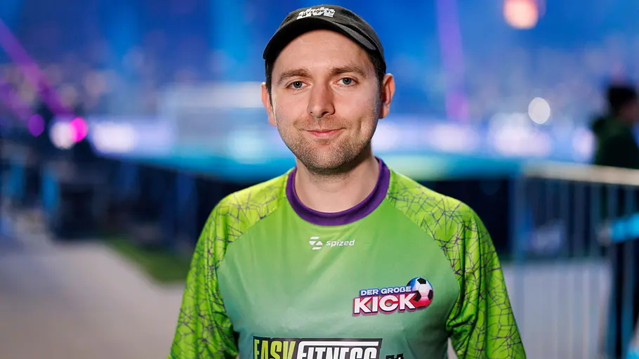 Adam Wolke gestorben Adam Wolke trägt ein grünes Sporttrikot und eine schwarze Kappe vor einer unscharfen, blau beleuchteten Veranstaltungskulisse.