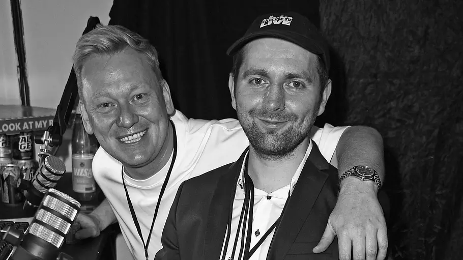 Adam Wolke Knossi Knossi legt links den Arm um den sitzenden Adam Wolke im Anzug mit Kappe. Neben ihnen stehen Mikrofone. Das Foto ist schwarz-weiß.