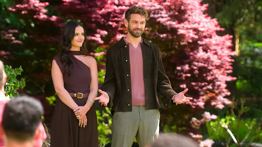 Age of Attraction Eine Frau in einem einseitigen, dunkelbraunen Kleid und ein Mann in rosa Shirt mit dunkler Jacke stehen in einem blühenden Garten vor einer Gruppe sitzender Personen. Im Hintergrund sind grüne Bäume und ein auffällig pink blühender Strauch zu sehen, während der Mann mit offenen Armen spricht.