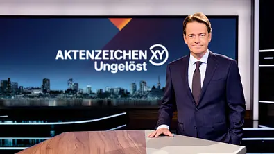 Rudi Cerne steht an einem Pult und moderiert „Aktenzeichen XY“. - Foto: ZDF/Nadine Rupp