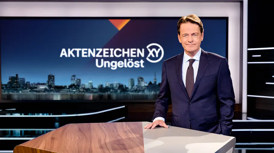 Rudi Cerne Rudi Cerne steht im dunklen Anzug hinter einem Studiopult. Im Hintergrund steht auf einem Bildschirm „Aktenzeichen XY Ungelöst“.