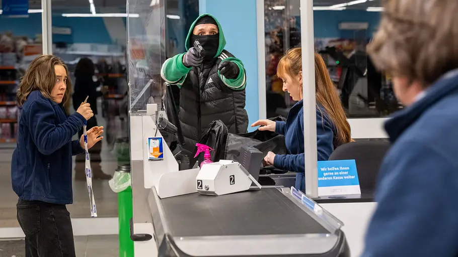 Maskierter Mann mit gezogener Pistole bedroht eine Kassiererin in einem Supermarkt, während eine Kundin erschrocken zurückweicht.