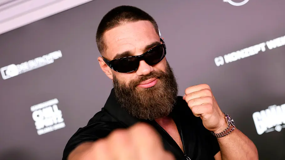 Aleks Petrovic Aleks Petrovic trägt Sonnenbrille und Vollbart und hält die Fäuste in Boxpose vor einer dunklen Wand mit Logos.