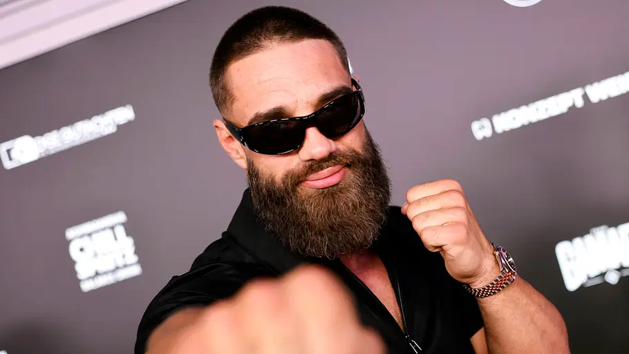 Aleks Petrovic will ins Dschungelcamp Aleks Petrovic trägt Sonnenbrille und Vollbart und hält vor einer Logowand die Fäuste hoch, eine davon nah an der Kamera.