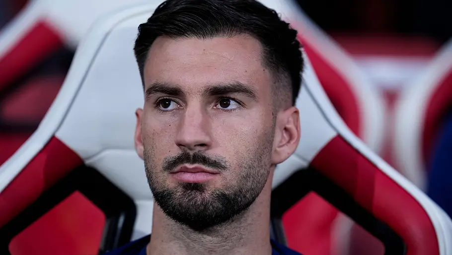 Ein Fußballspieler sitzt mit ernstem Blick auf der Ersatzbank in einem rot-weiß gepolsterten Stadionsitz und verfolgt aufmerksam das Spielgeschehen.