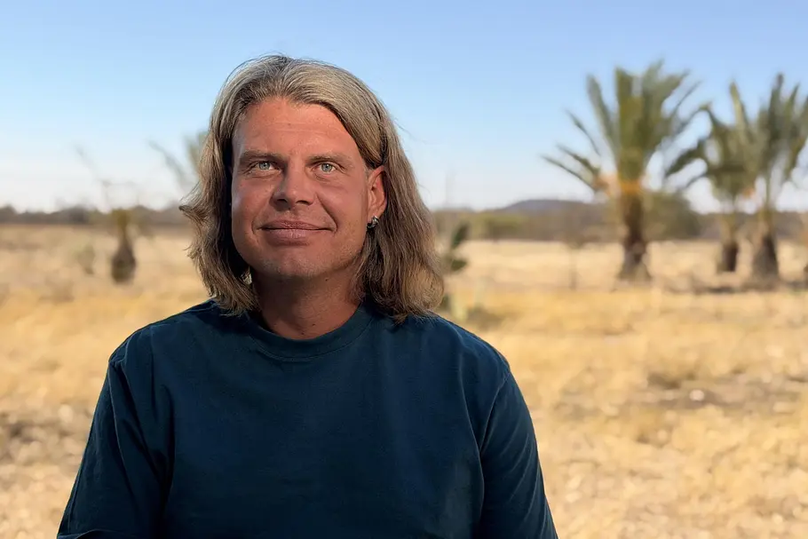 Alex mit langen Haaren entspannt vor Palmen.
