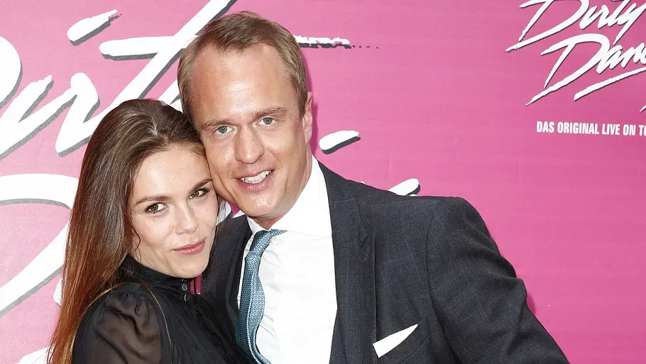 Alexander und Angelina Posth Alexander Posth hält das angewinkelte Bein von Angelina Posth im schwarzen Kleid. Sie posieren vor einer pinken „Dirty Dancing“-Wand.