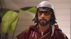 Ali G trägt eine weiße Kappe, eine gelb getönte Brille, eine glänzende bordeauxrote Kapuzenjacke und eine goldene Kette. - Foto: TMDB Ali G trägt eine weiße Kappe, eine gelb getönte Brille, eine glänzende bordeauxrote Kapuzenjacke und eine goldene Kette. - Foto: TMDB